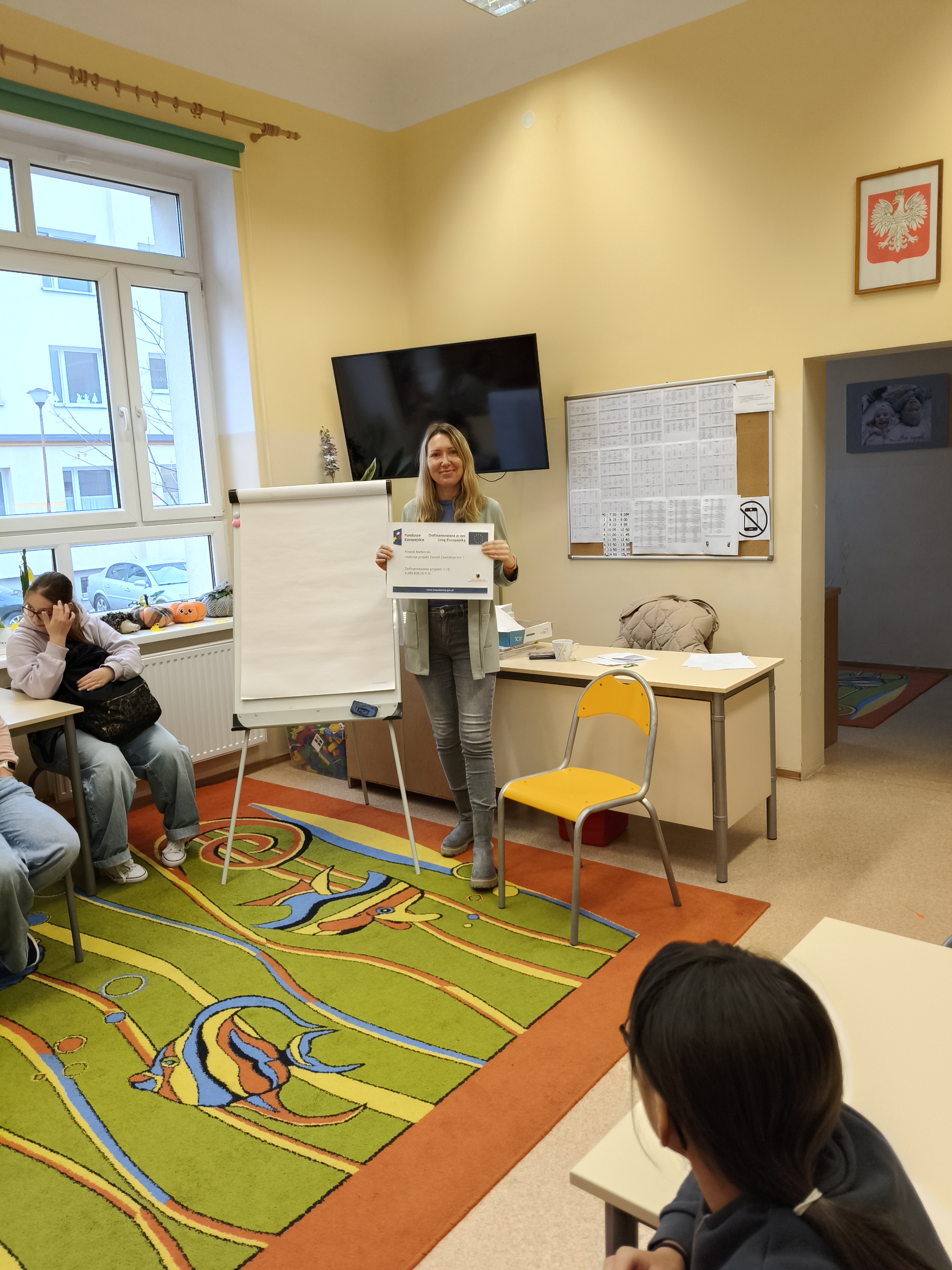 Zdjęcie przedstawia Koordynatorkę Powiatową stojącą przy tablicy w świetlicy szkolnej w otoczeniu uczniów i trzymającą planszę z informacją o projekcie "Zostań Zawodowcem 2" 