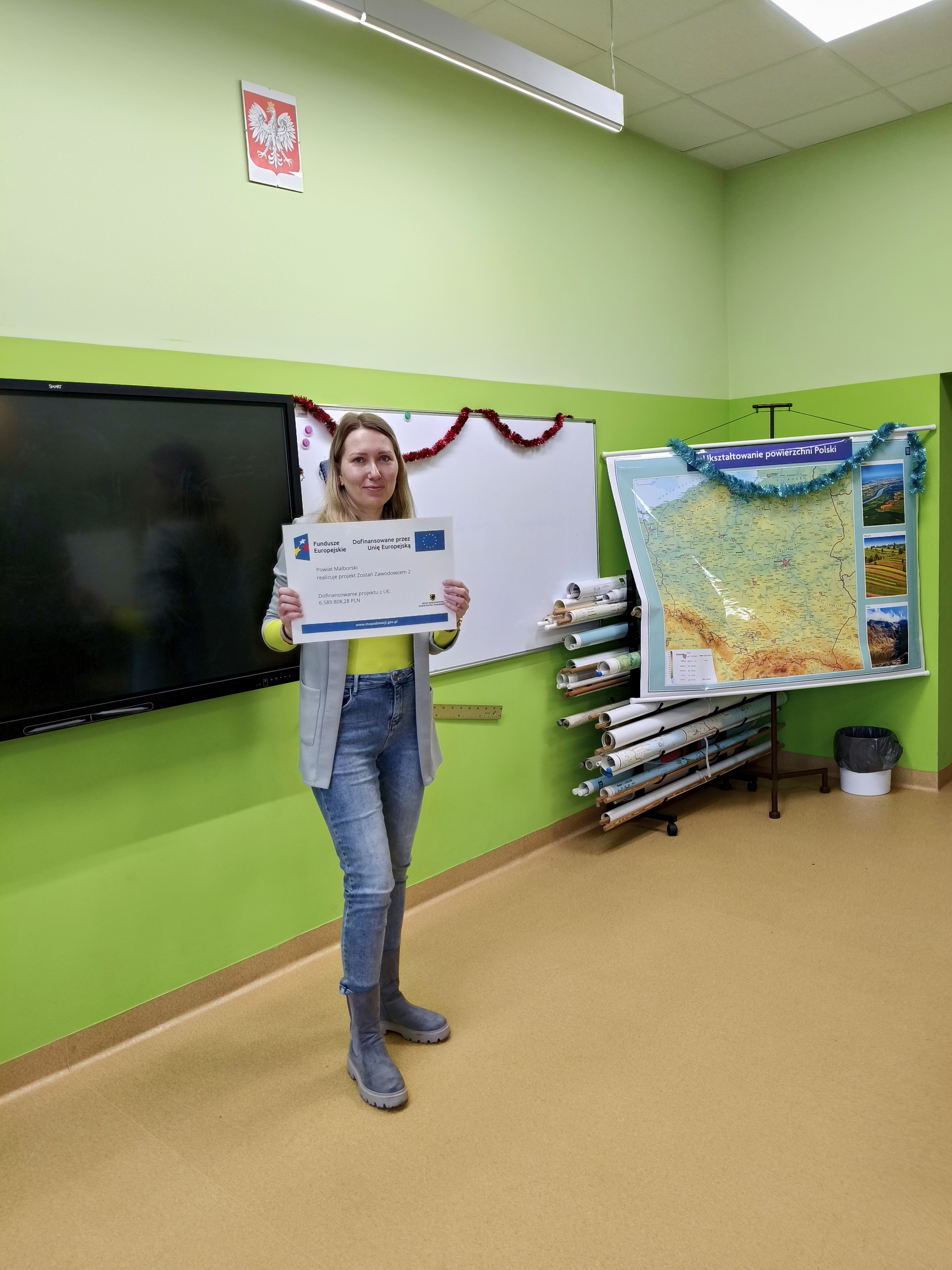 Zdjęcie przedstawia Koordynatorkę Powiatową stojącą w klasie na tle tablicy, prezentującą planszę z informacją o realizowanym projekcie "Zostań Zawodowcem 2" 