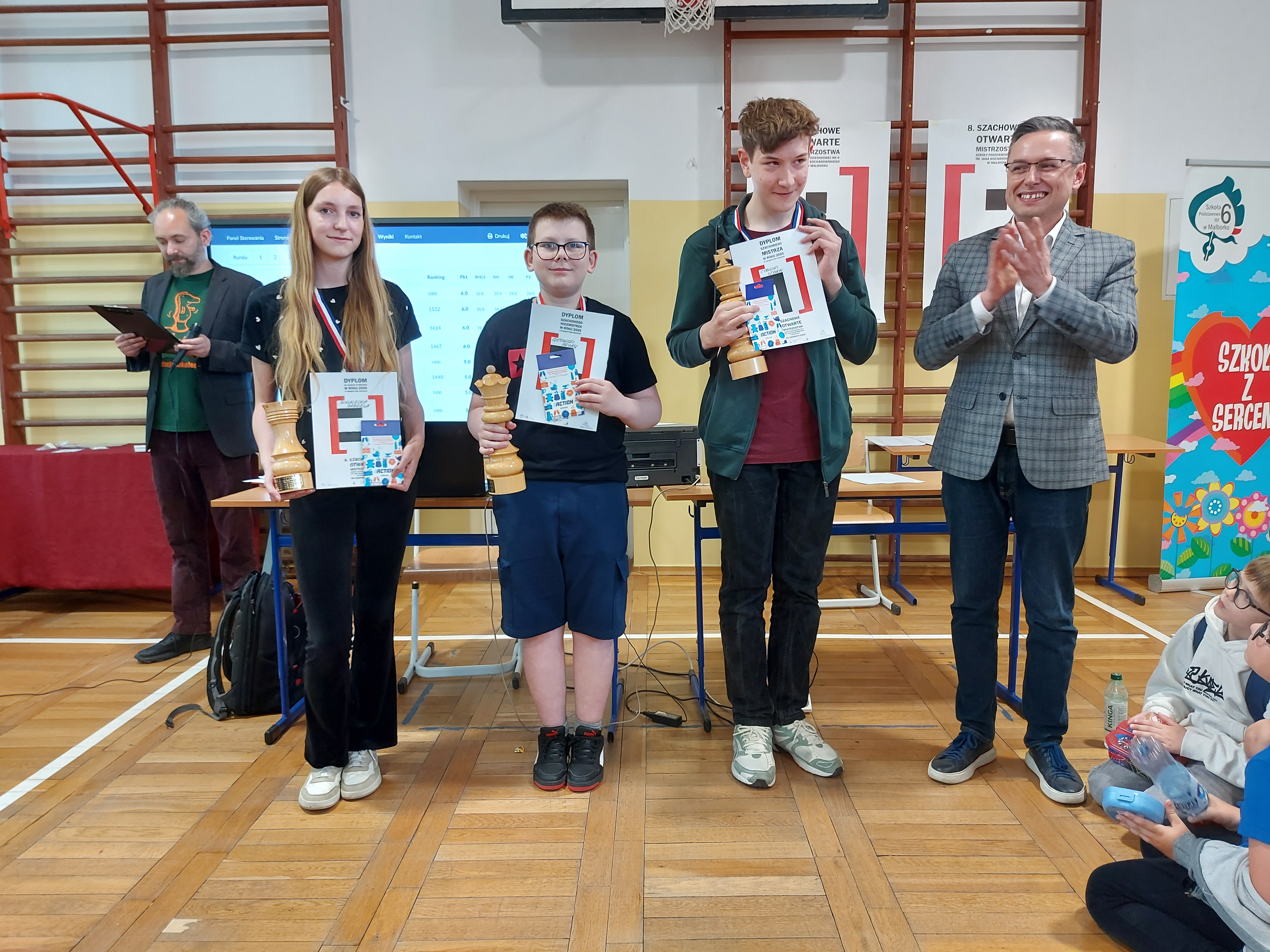 Zdjęcie przedstawia zwycięskich uczniów trzymających dyplomy w towarzystwie prowadzącego wydarzenie oraz dyrektora szkoły